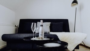 Un dormitorio moderno con un sofá negro, una pequeña mesa redonda con una botella y vasos, y un suave plaid blanco sobre el sofá.