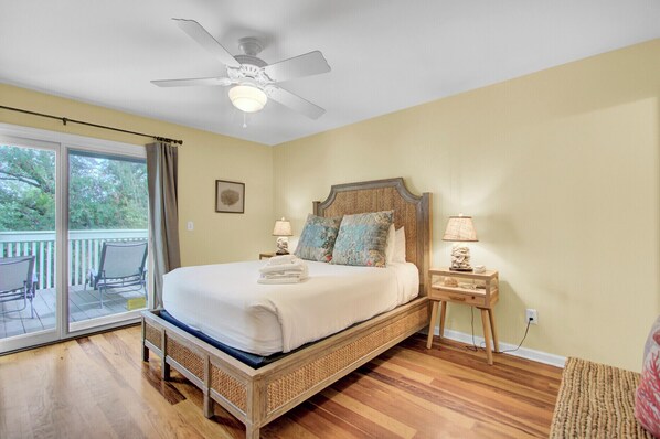 A bedroom with a large bed, a ceiling fan, a nightstand, and a view of the outdoors.