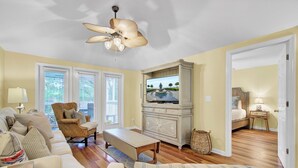 A living room with a television, a ceiling fan, a wicker chair, a sofa, and a bed in the adjacent room. - Award-Winning Resort Amenities | Sandpiper Court 14 (Isle of Palms)