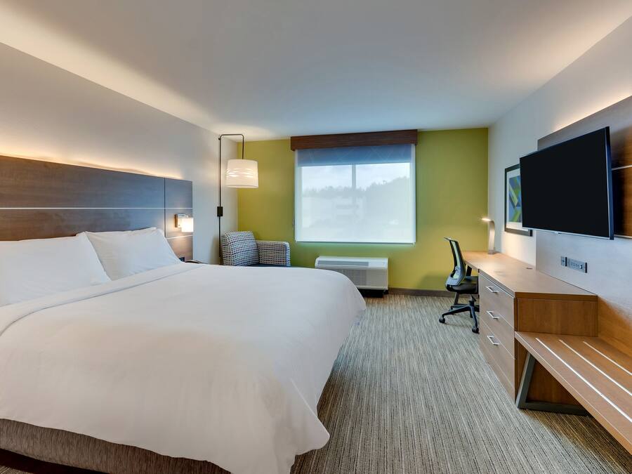 A hotel room with a large bed, a desk with drawers, a flat-screen TV, a window with a view, and a wall-mounted light fixture.