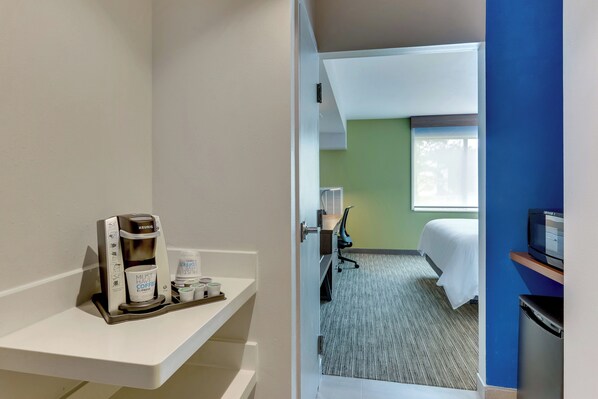A hotel room with a coffee maker on a white shelf, a bed with white linens, a desk with a chair, and a television on a stand.