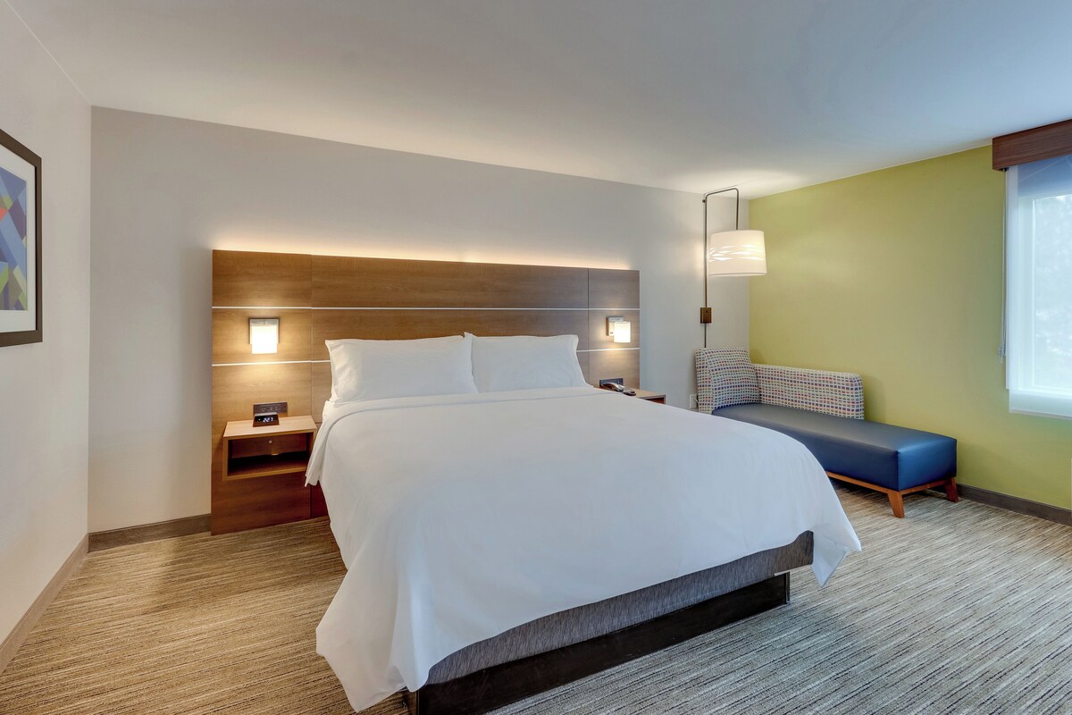 A modern hotel room with a large bed, bedside tables, a nightstand, a bench, and a wall-mounted lamp.