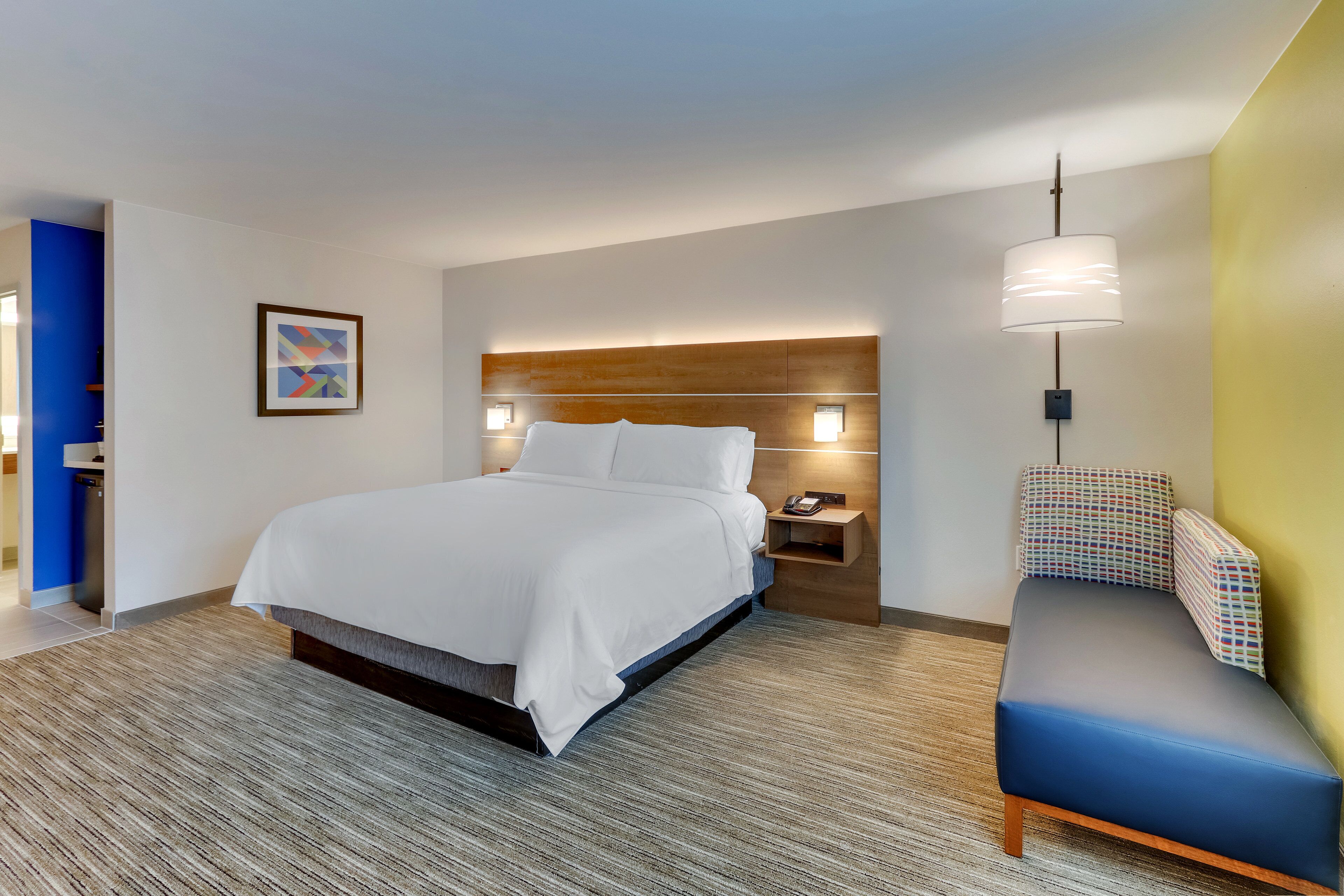 A modern hotel room with a large bed, bedside tables, a sofa, and a wall-mounted lamp.