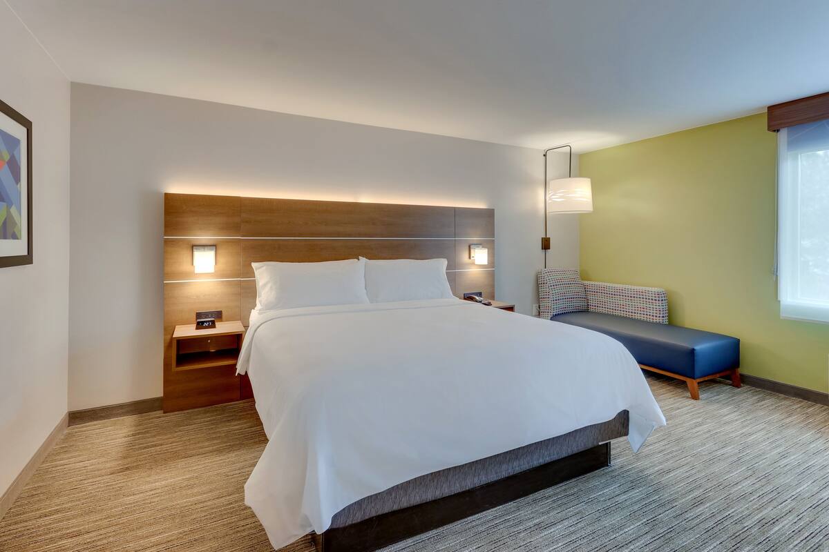 A modern hotel room with a large bed, bedside tables, a nightstand, a bench, and a wall-mounted lamp.