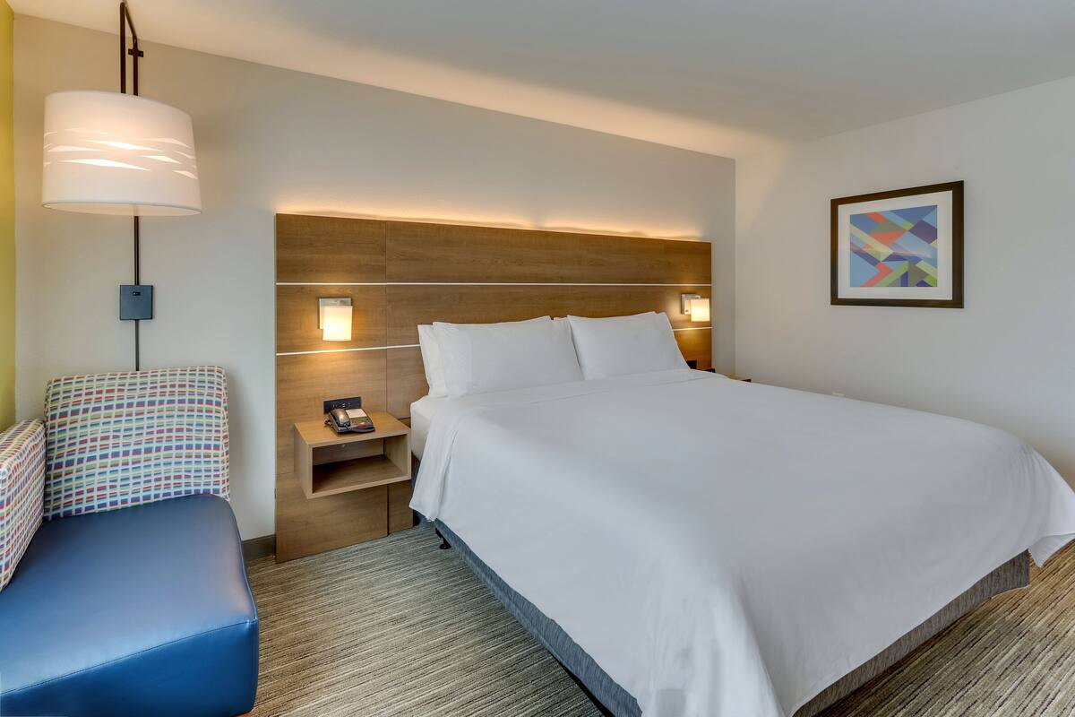 A hotel room with a large bed, a bedside table, a lamp, a chair, and a framed picture on the wall.