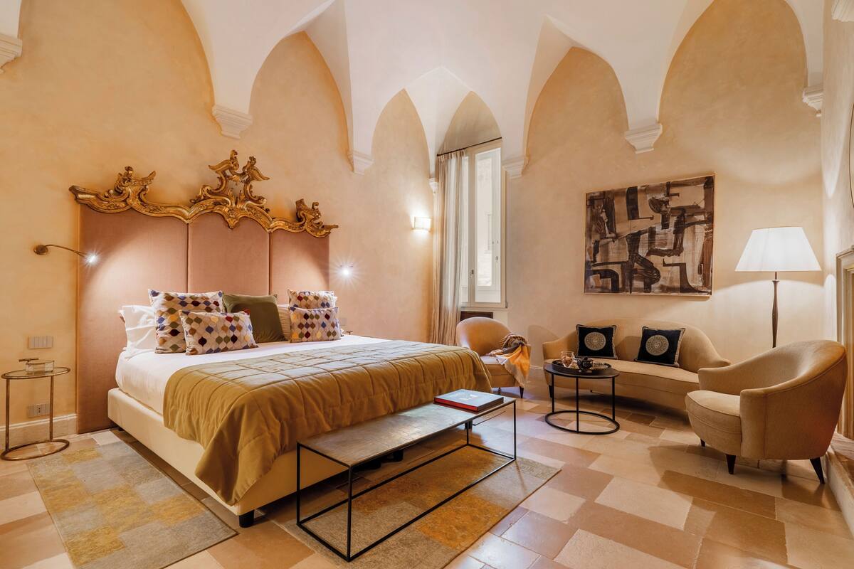 A bedroom with a large bed, a bedside table, a chair, a sofa, and a painting on the wall.