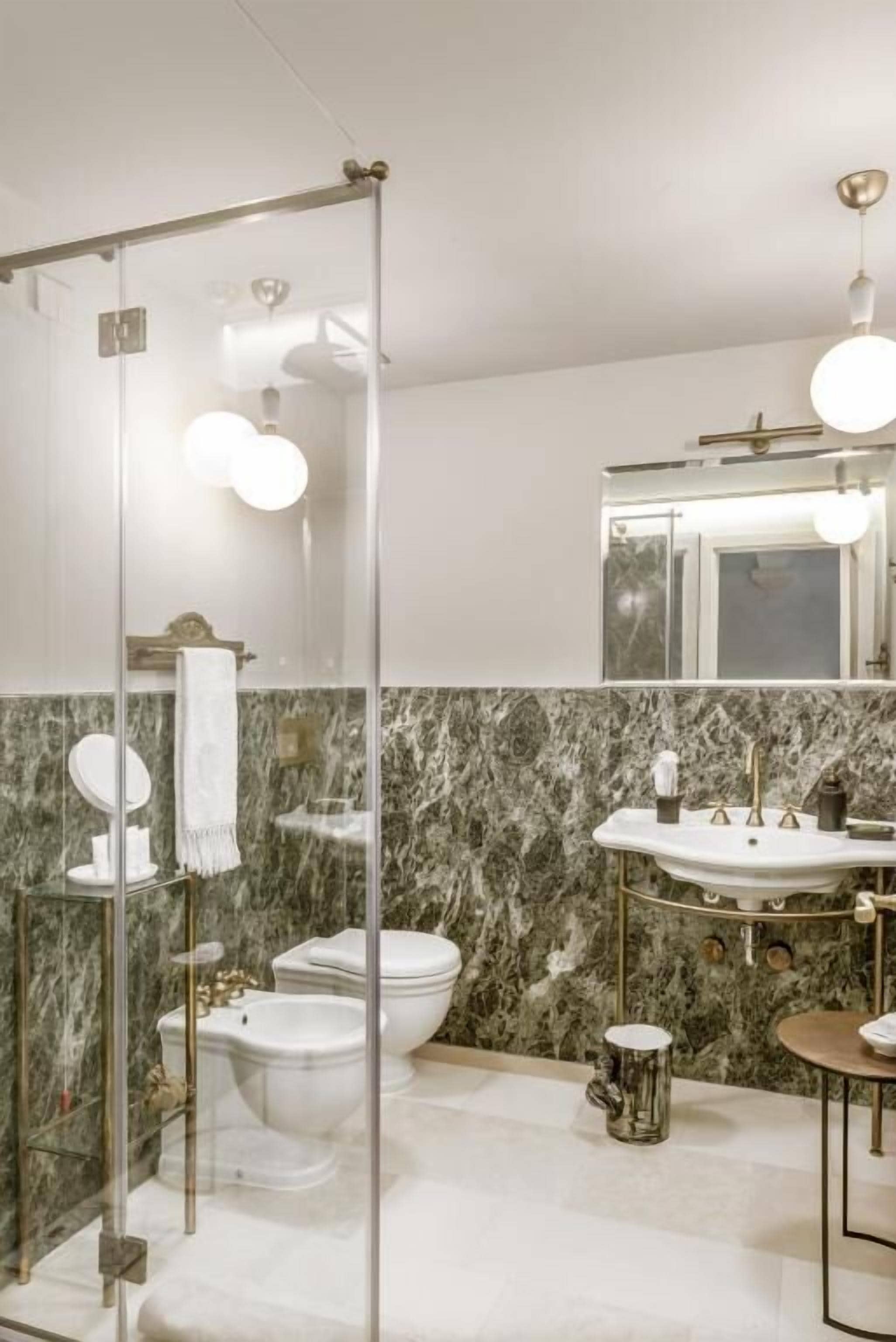 A modern bathroom with a glass shower enclosure, a marble-tiled wall, a white sink with gold fixtures, and a large mirror above the sink.