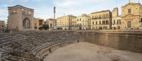 An ancient amphitheater with tiered seating, a central stage, and a backdrop of historic buildings.
