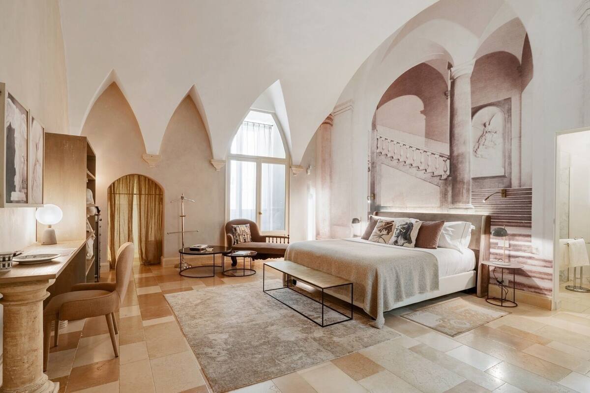 A spacious bedroom with a large bed, a desk, and a chair. There are arched ceilings and a view of a staircase.
