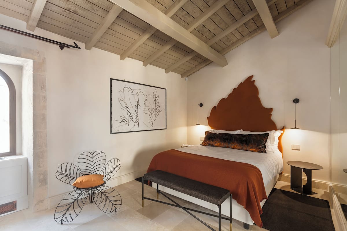 A bedroom with a bed, a bench, a chair, a framed artwork, and a unique headboard.