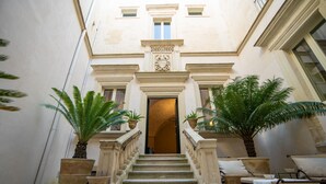 A grand staircase leading to a door with a decorative emblem, flanked by potted plants and a bench. - Palazzo Maresgallo (Lecce)