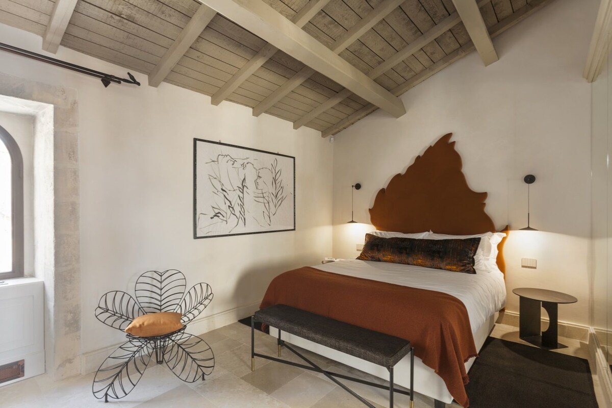 A bedroom with a bed, a bench, a wall-mounted light, and a framed artwork.