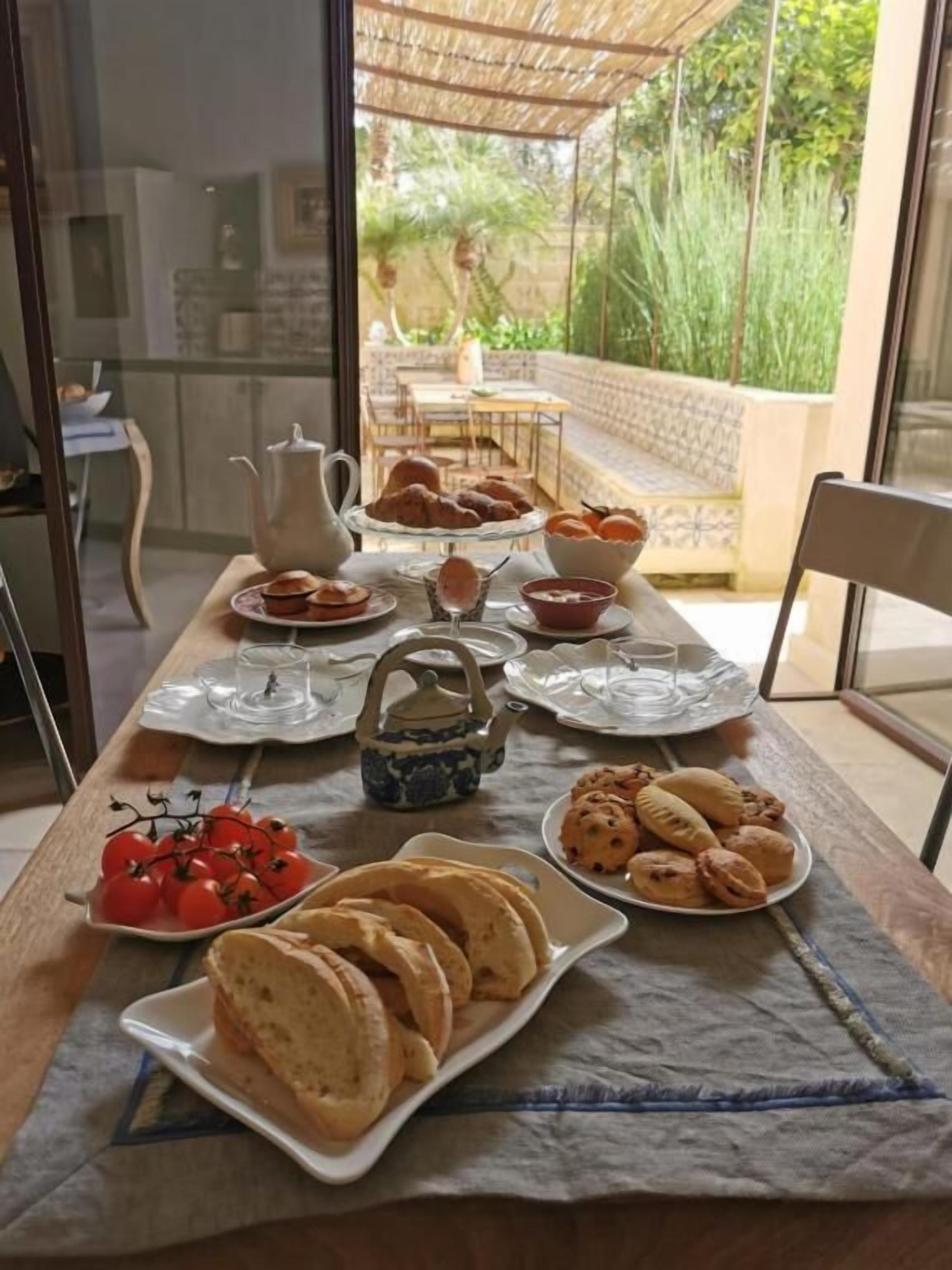 A breakfast table with various pastries, bread, and a teapot, set in a room with a view of a garden and outdoor seating.