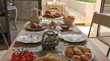 A breakfast table with various pastries, bread, and a teapot, set in a room with a view of a garden and outdoor seating.