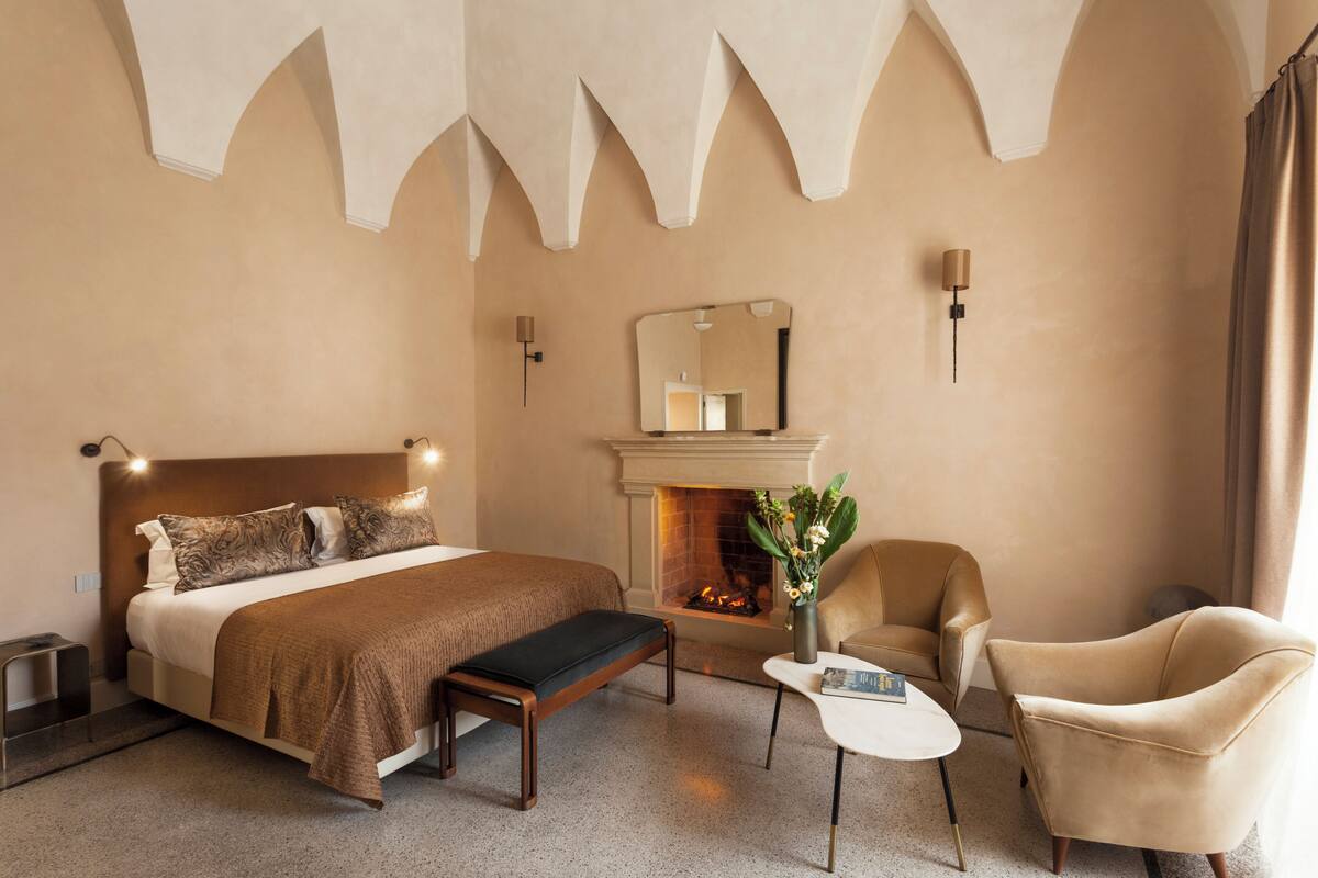 A hotel room with a large bed, a bench, a small table, two armchairs, a fireplace, and a mirror on the wall.