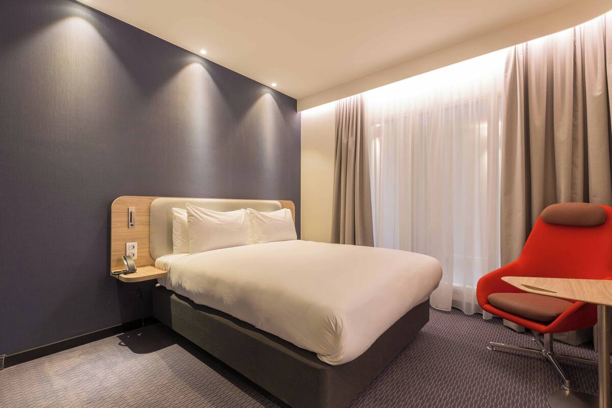 A hotel room with a bed, a nightstand, a red chair, and a window with curtains.