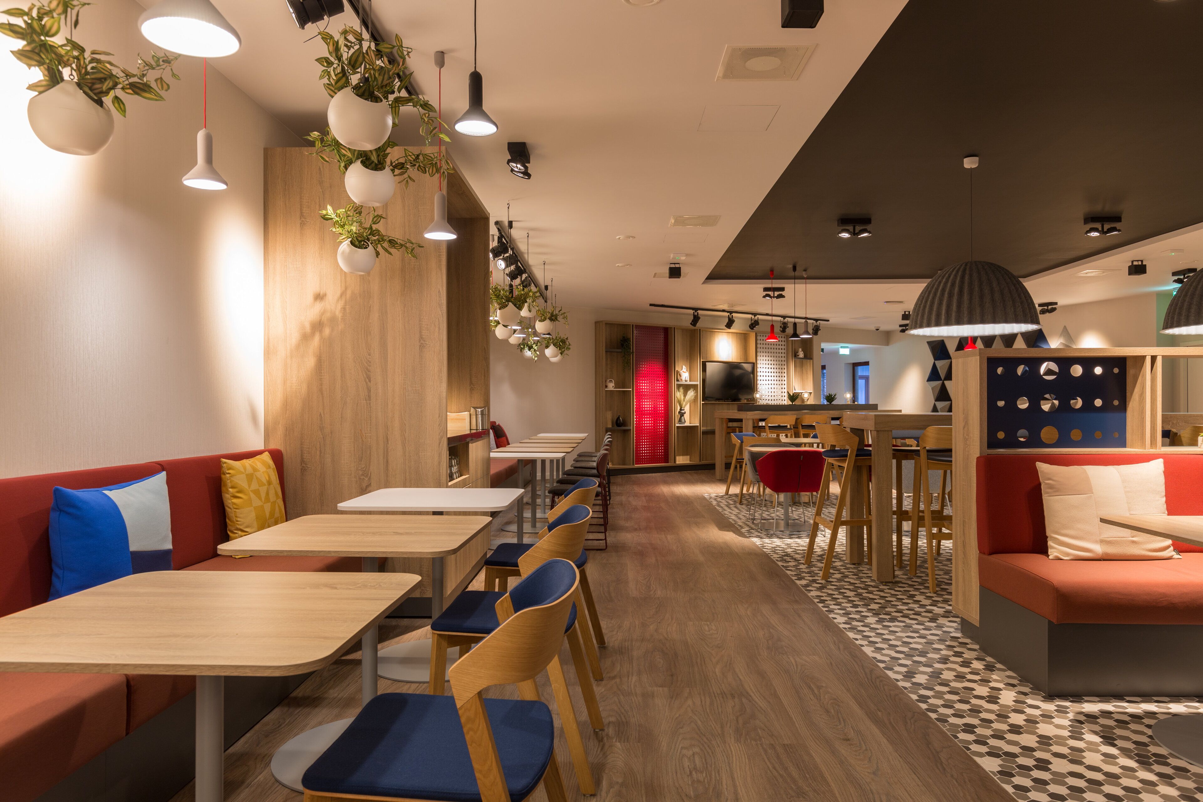 A modern restaurant interior with wooden tables, red and blue upholstered seating, hanging plants, and pendant lighting.