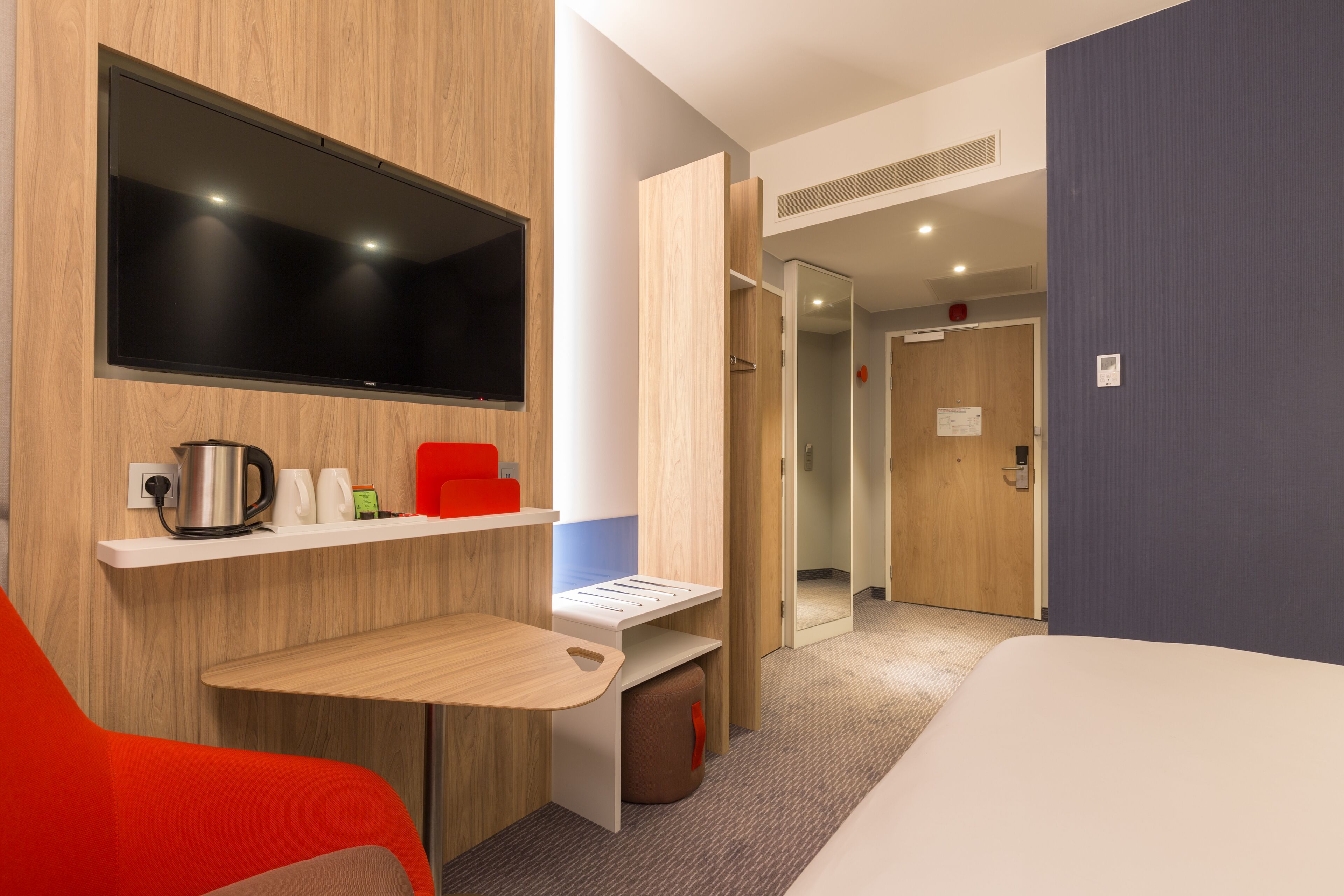 Una habitación de hotel moderna con televisión de pantalla plana, un escritorio de madera, una silla roja, una cama y un pasillo que conduce a otra habitación.