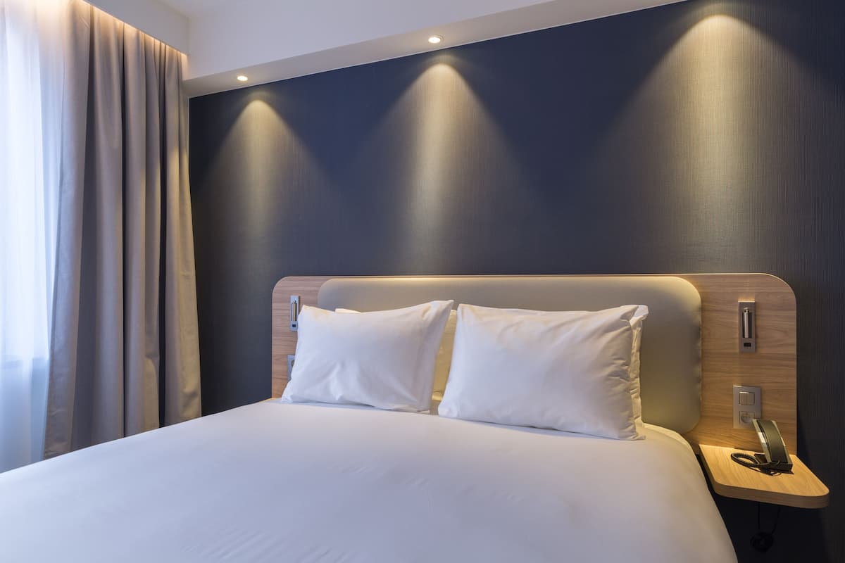 A hotel room with a large bed, two pillows, a bedside table with a phone, and a wall-mounted light.