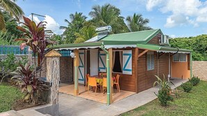 Una pequeña casa de madera con techo verde, un porche cubierto con mesa y sillas, y un jardín con plantas tropicales.