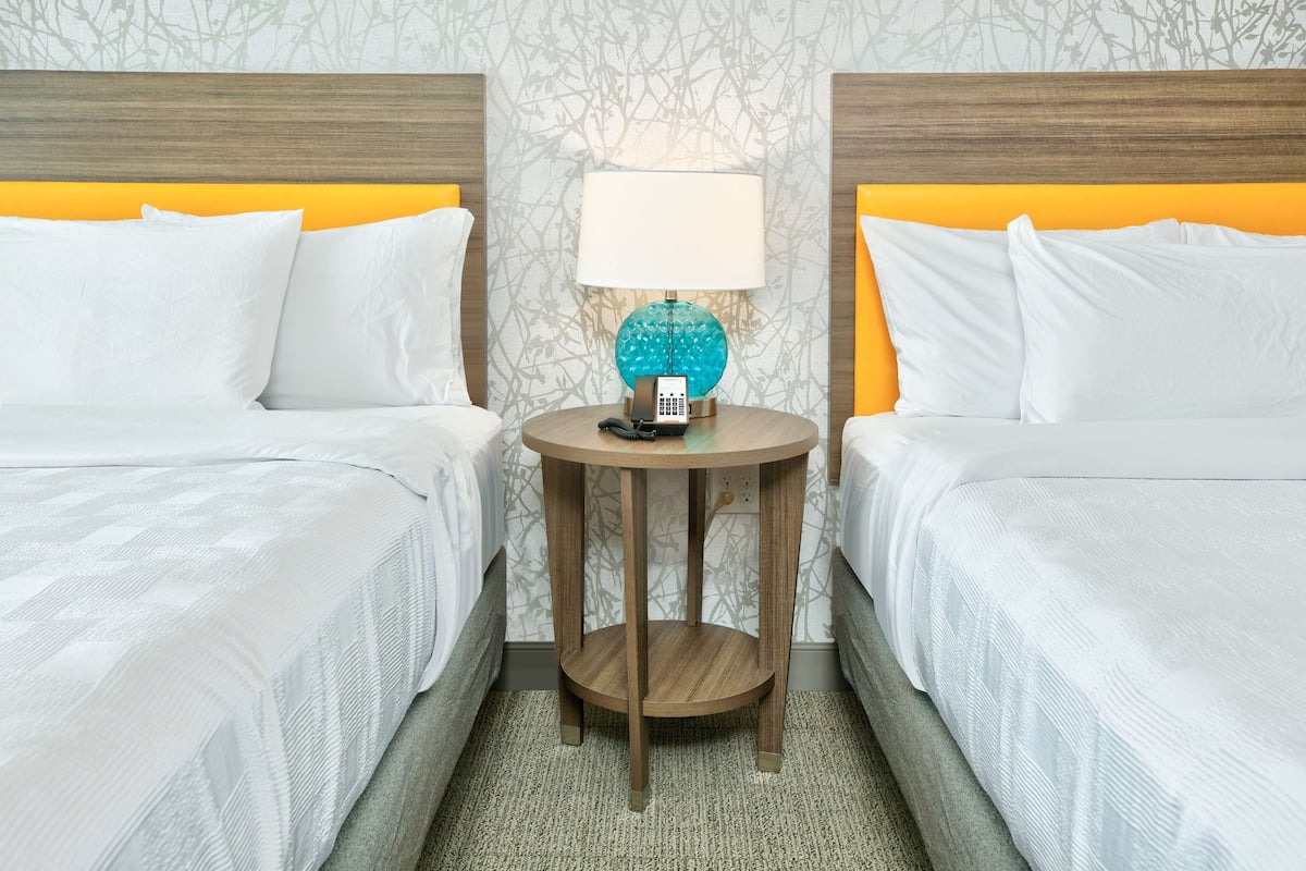 A hotel room with two beds, a round wooden nightstand with a lamp, and a patterned wallpaper.