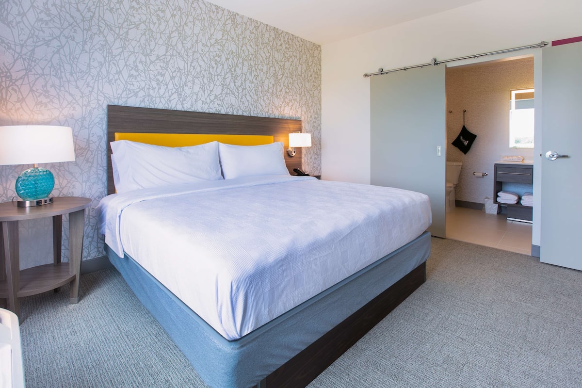 A modern hotel room with a large bed, two bedside tables, a nightstand, and a bathroom visible through an open door.