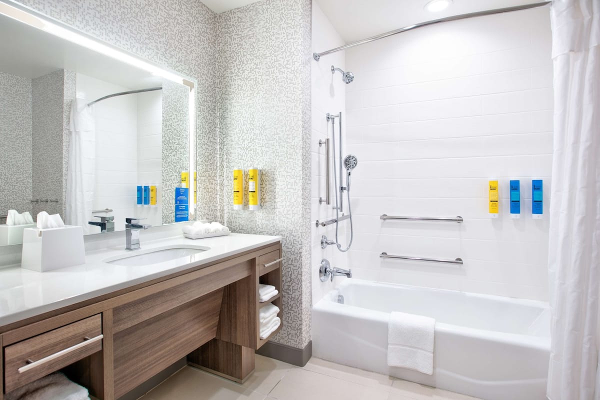 A modern bathroom with a large mirror, two sinks, a bathtub, and toiletries on the counter.