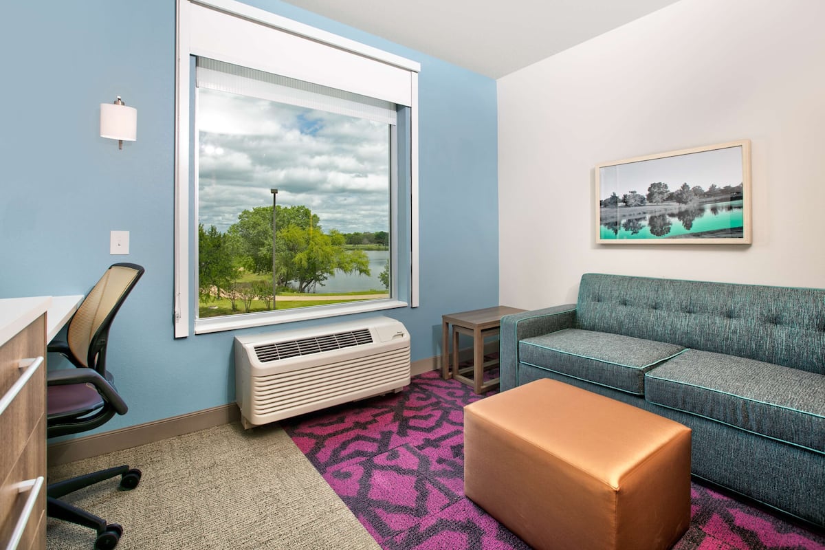A room with a large window, a blue wall, a grey sofa, a brown ottoman, and a small table.