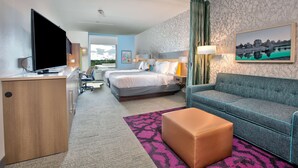 A hotel room with a bed, a sofa, a desk with a TV, a desk lamp, a painting, and a view of the outdoors. - Home2 Suites by Hilton Wichita Falls, TX (Wichita Falls)