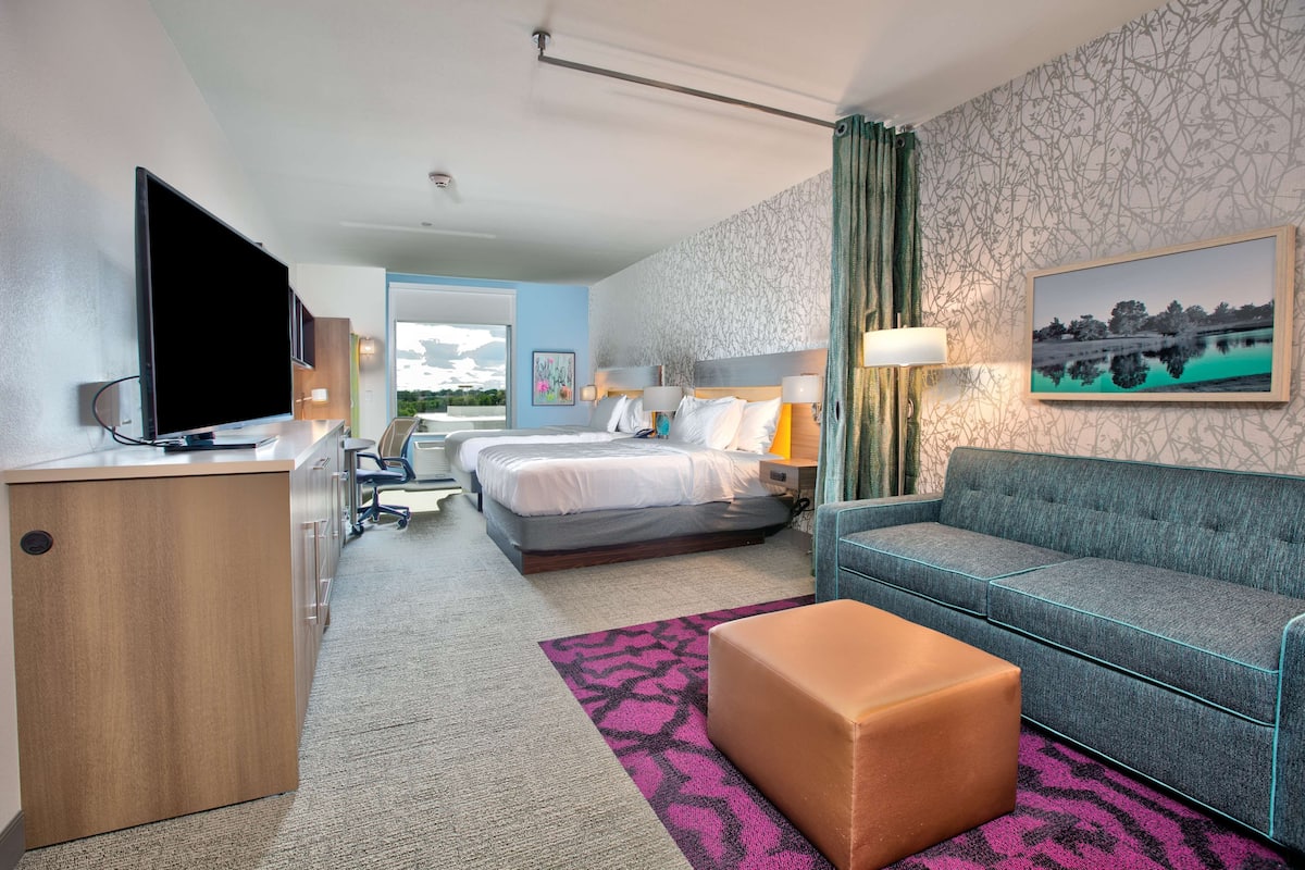 A hotel room with a bed, a sofa, a desk with a TV, a desk lamp, a painting, and a view of the outdoors.