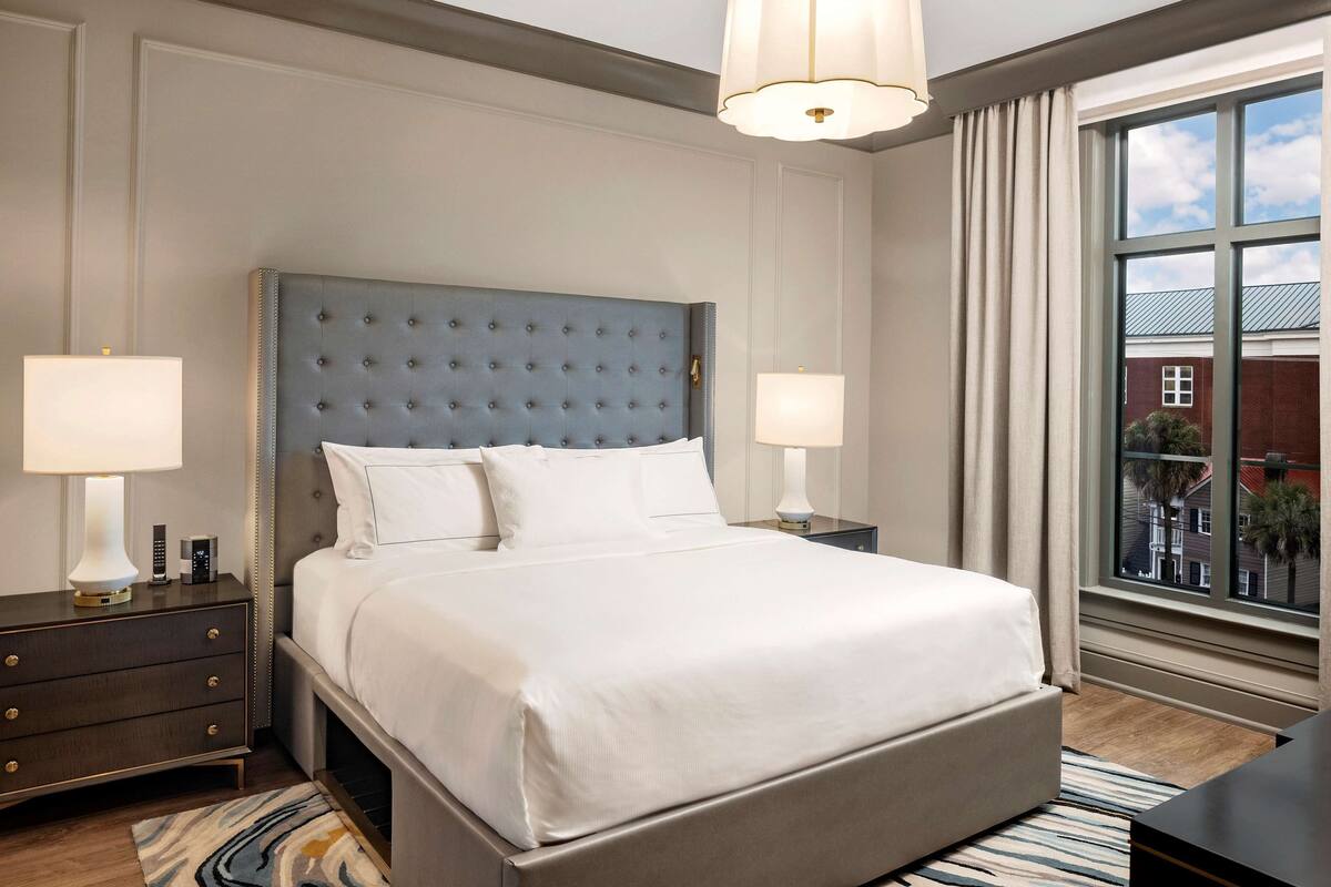 A neatly made bed with a tufted headboard, two bedside lamps, a dark wooden nightstand, and a view of the outside through a large window.