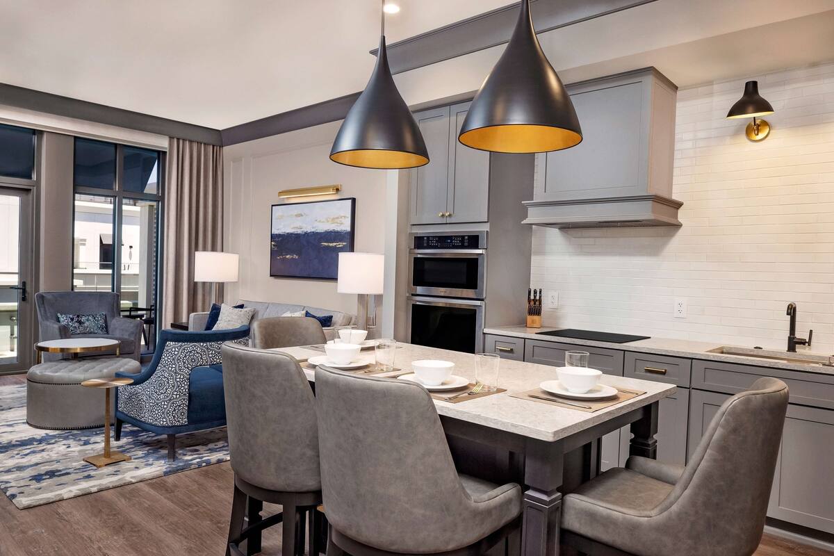 A modern kitchen with grey cabinetry, a central island with bar stools, pendant lighting, and a view of the city through large windows.