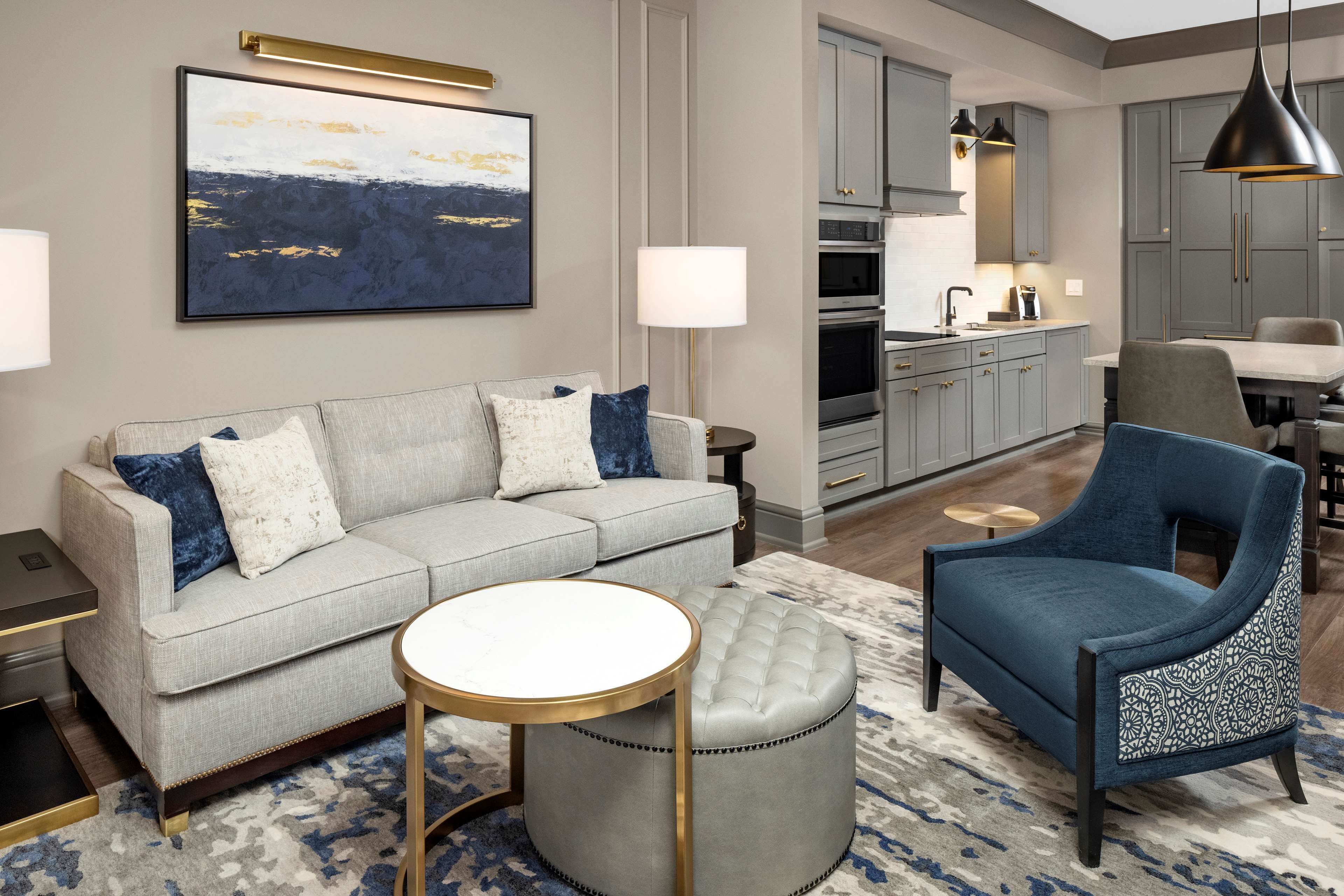 Un salon moderne avec un canapé gris, un fauteuil bleu, une table basse ronde et un pouf à motifs.