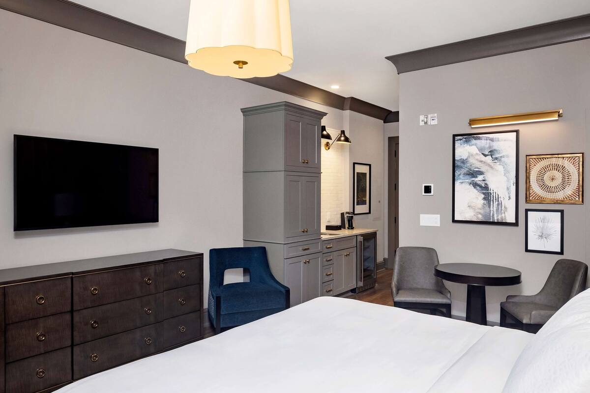 A modern hotel room with a large bed, a TV mounted on the wall, a gray wardrobe, a blue chair, a small table, and artwork on the walls.