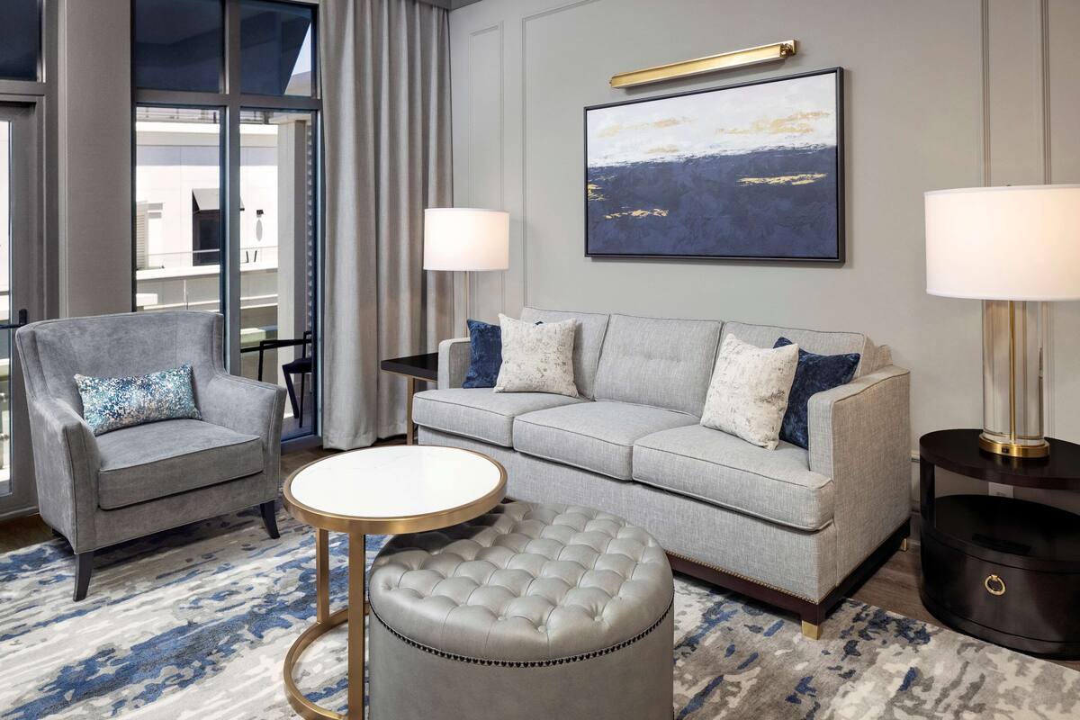 A modern living room with a grey sofa, two armchairs, a round coffee table, and a tufted ottoman. There are two floor lamps and a large abstract painting on the wall.