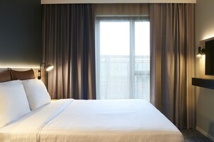 A hotel room with a large bed, a TV mounted on the wall, and a view of the city through the window.