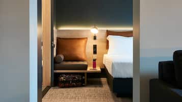 A compact hotel room with a bed, a sofa, a nightstand with a lamp, and a suitcase on the floor.