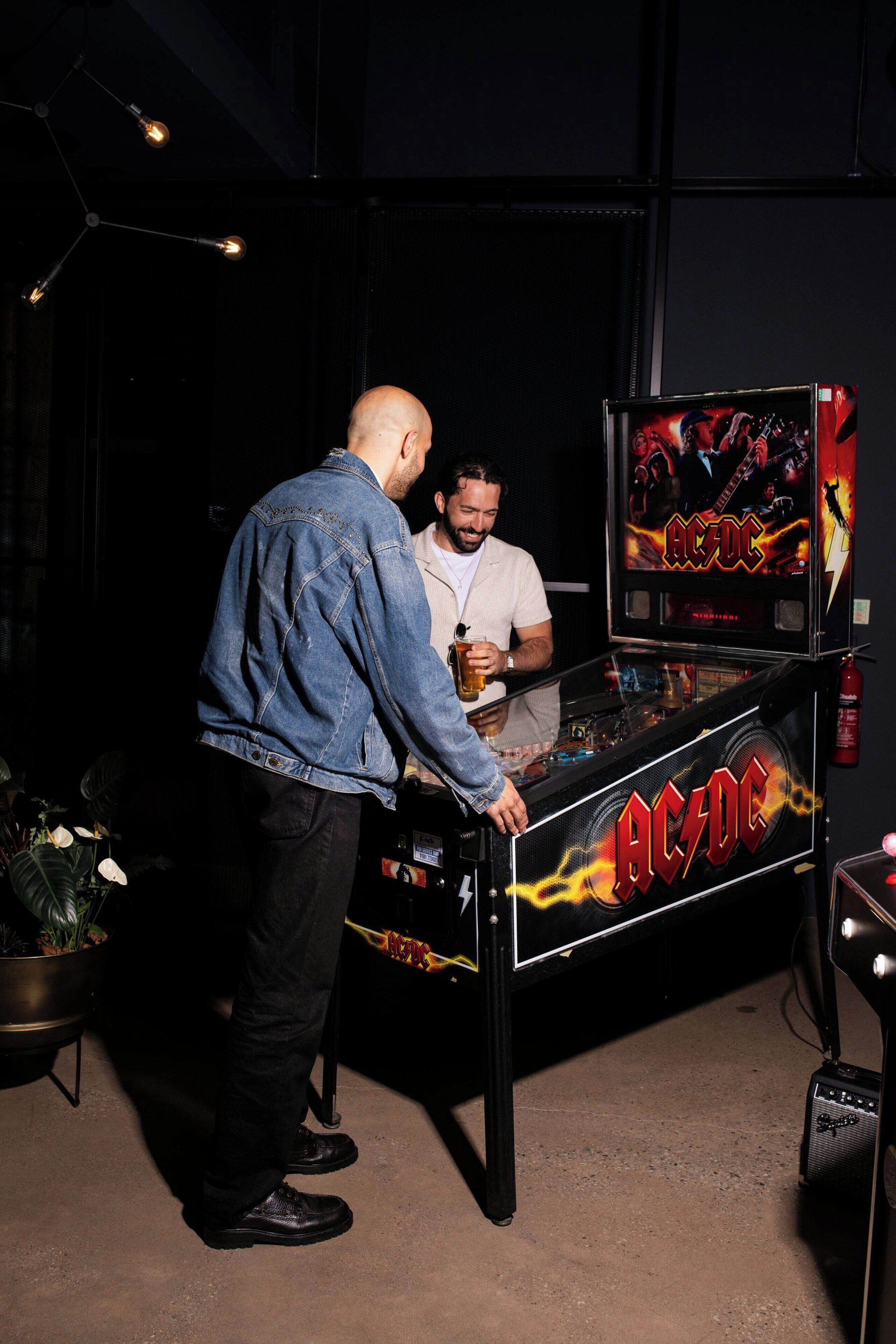 A man standing at a pinball machine with a drink in hand.