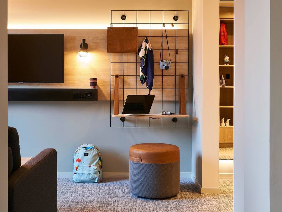 A modern living room with a flat-screen TV, a laptop on a wooden shelf, a round ottoman, and a wall-mounted storage unit with hanging items.