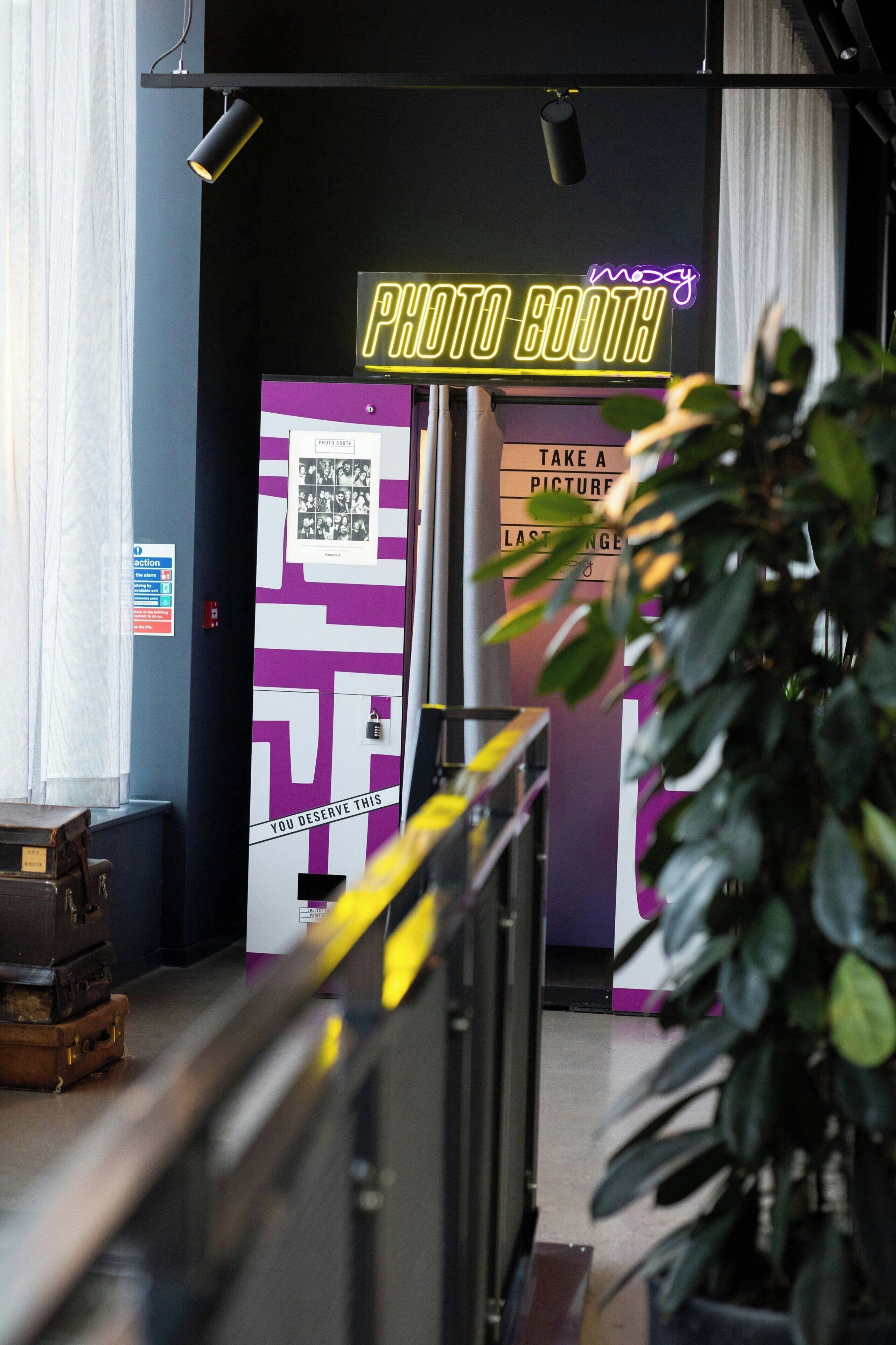 Uma cabine de fotos com um letreiro neon, um código QR e uma placa que diz 'Tire uma foto com duração de 8 segundos'.