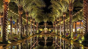 A nighttime view of a hotel with palm trees lined up along a reflective pool, leading to a building with a fountain in the center.