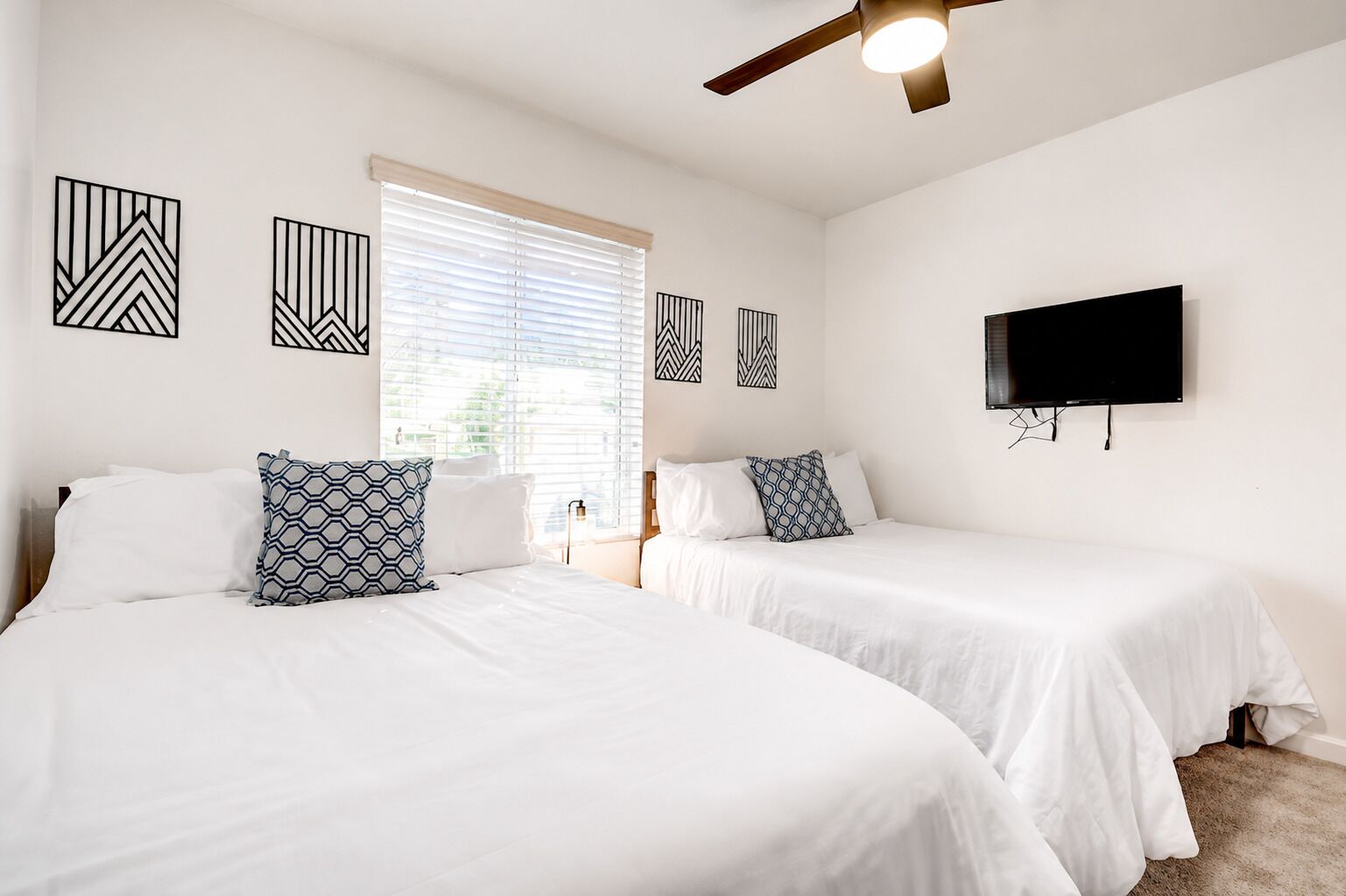 A hotel room with two beds, a ceiling fan, a TV, and artwork on the walls.