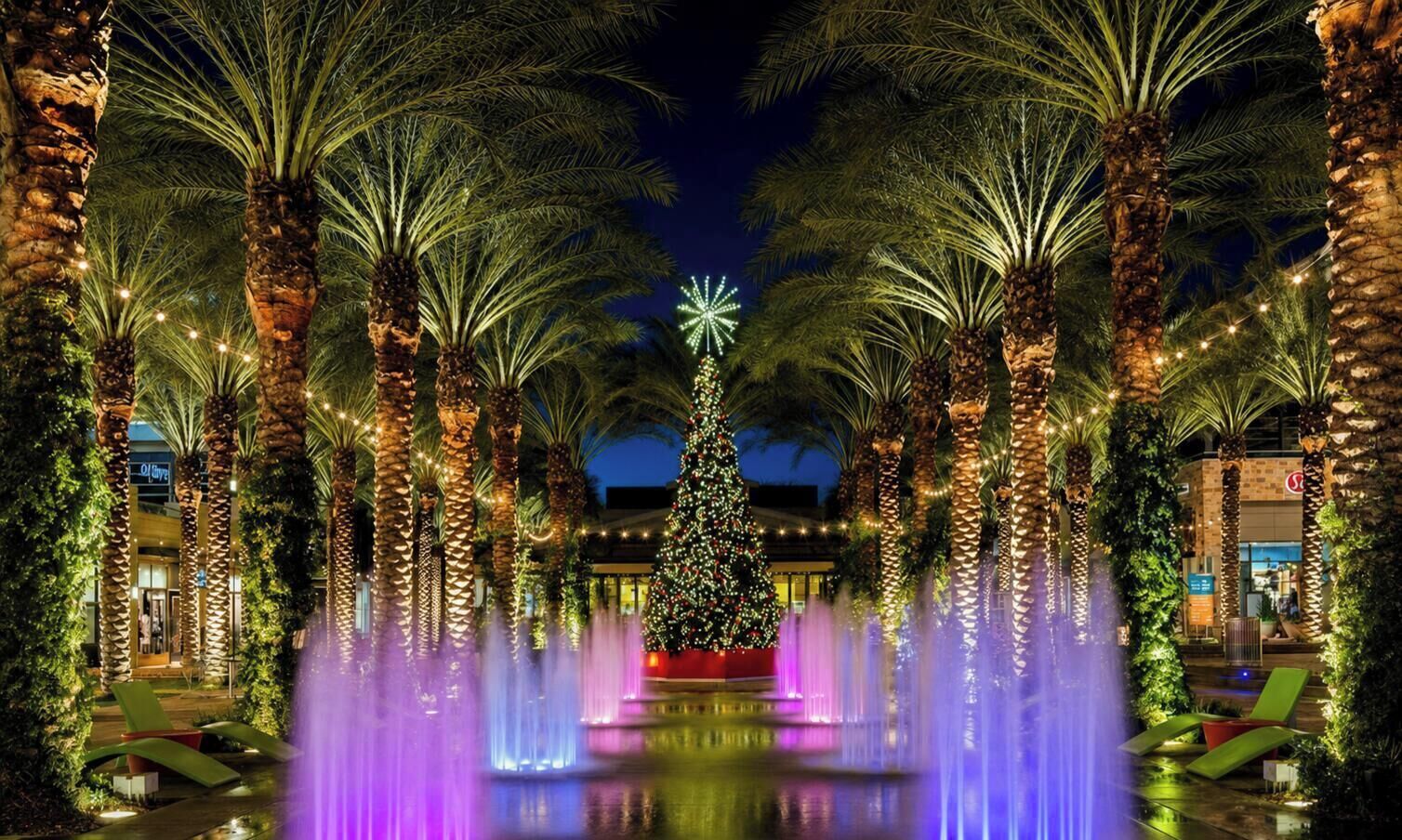 Een nachtscène met een versierde kerstboom en palmbomen, omgeven door waterpartijen en verlicht met lampjes.