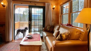 Een gezellig chaletinterieur met een houten plafond, een bank, een bijzettafeltje en een hond die bij een schuifpui staat.
