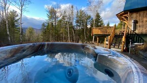 Een hottub in een bebost gebied, met een houten terras en een gebouw op de achtergrond.