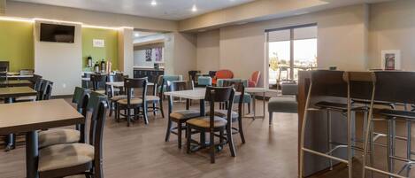 A modern dining area with multiple tables and chairs, a kitchen area in the background, and a large window providing natural light.