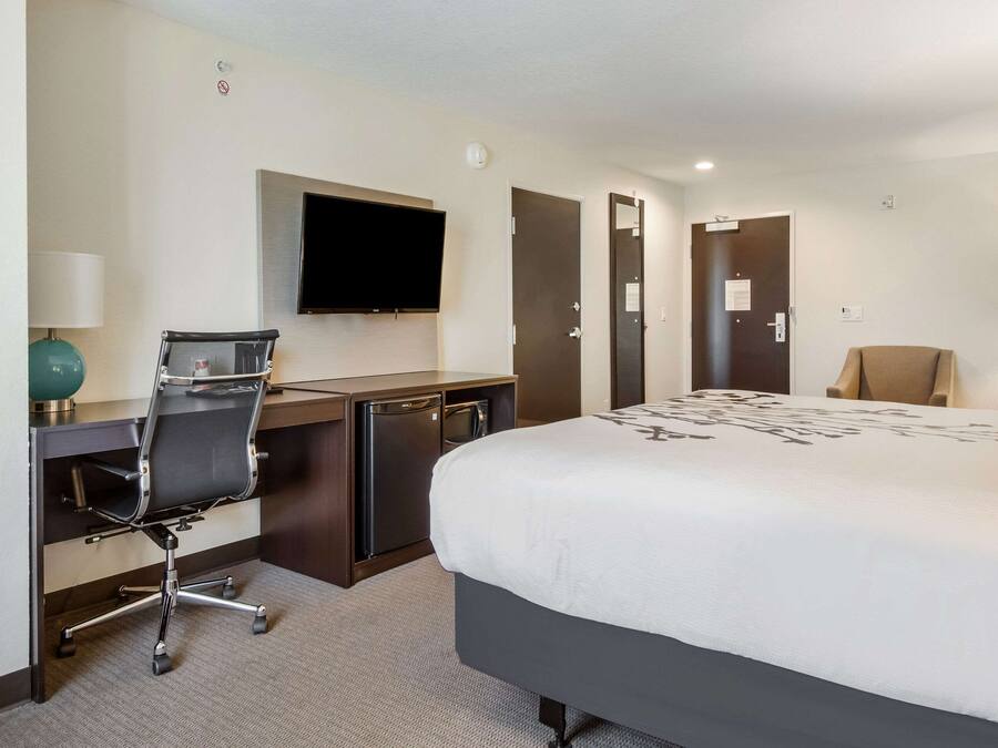Quarto de hotel com cama, escrivaninha, cadeira, televisão e uma pequena cozinha.
