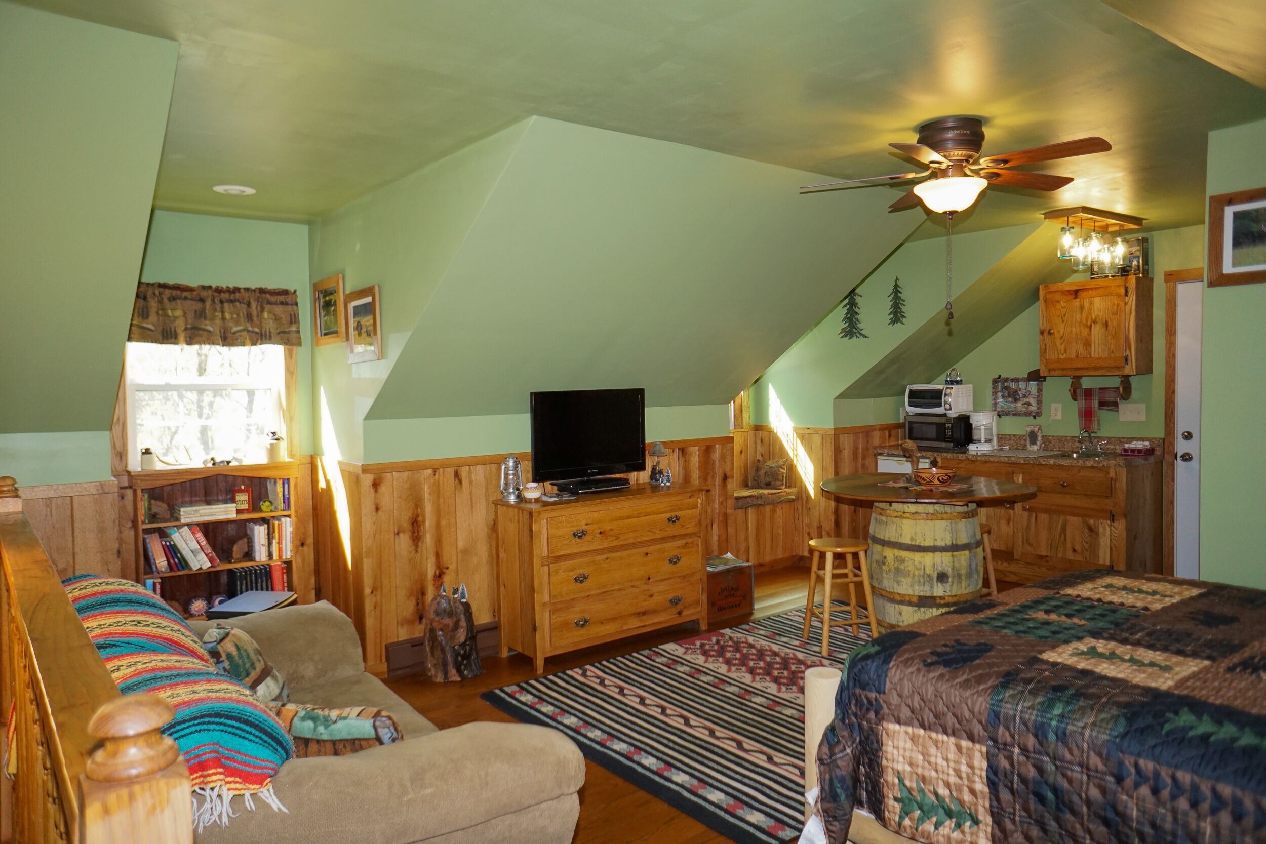 A cozy attic bedroom with a sloped ceiling, a ceiling fan, a television, a wooden desk, a kitchen area, and a large bed with a patterned bedspread.