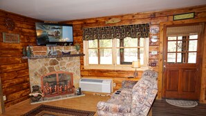 A cozy cabin interior with a stone fireplace, a television mounted on the wall, a patterned sofa, and a wooden door. - King's River Cabin-River Frontage - Fire Pit - Screened Porch (Bryson City)