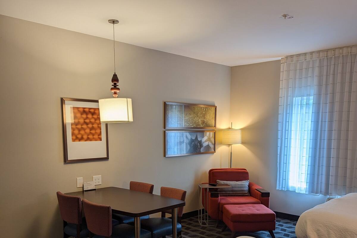 Habitación de hotel con cama, escritorio con sillas, sillón rojo, lámpara y dos cuadros en la pared.